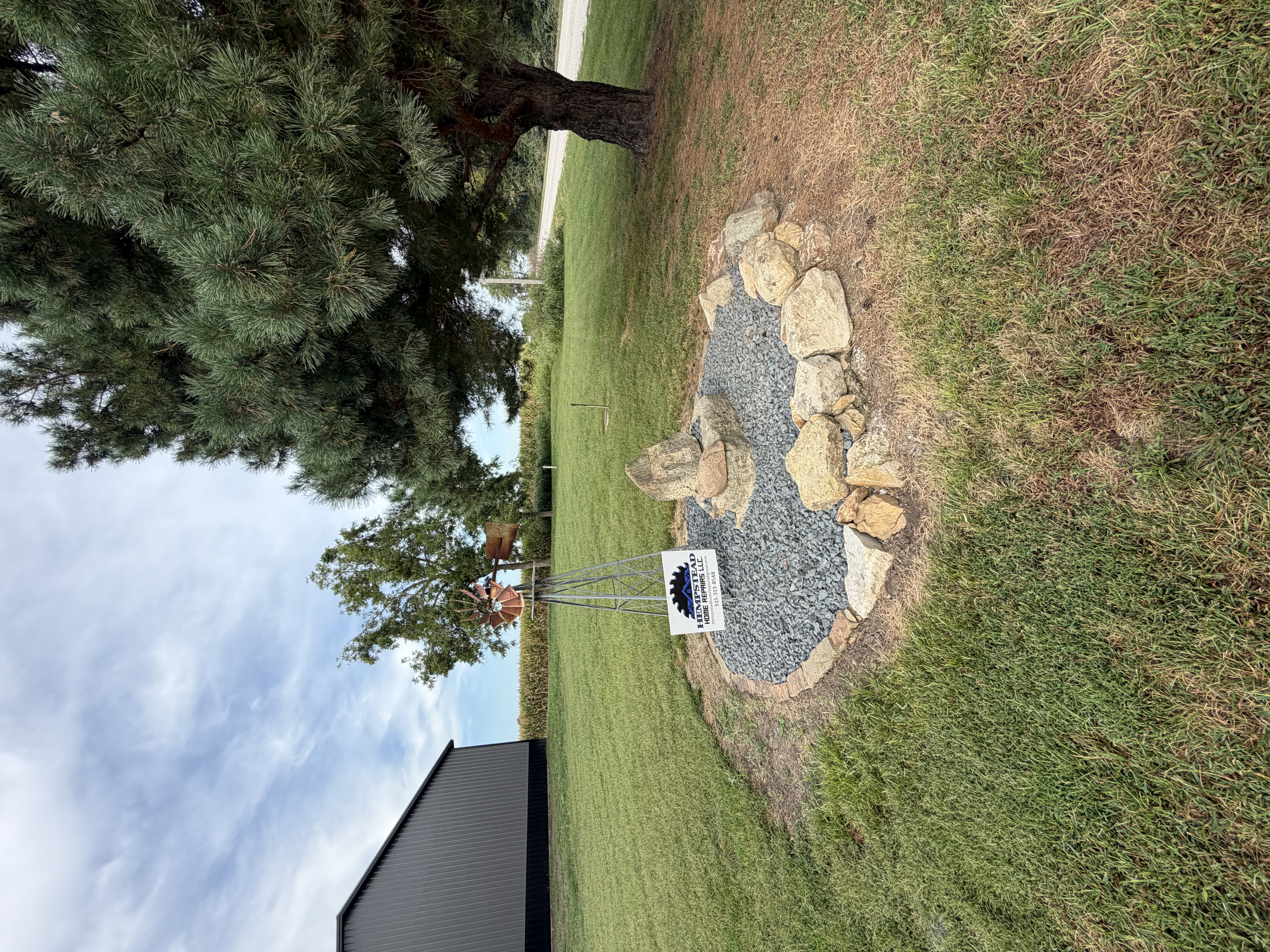 A landscaped garden bed filled with grey crushed stone and bordered by large natural tan rocks, featuring a small metal windmill on a grassy lawn.