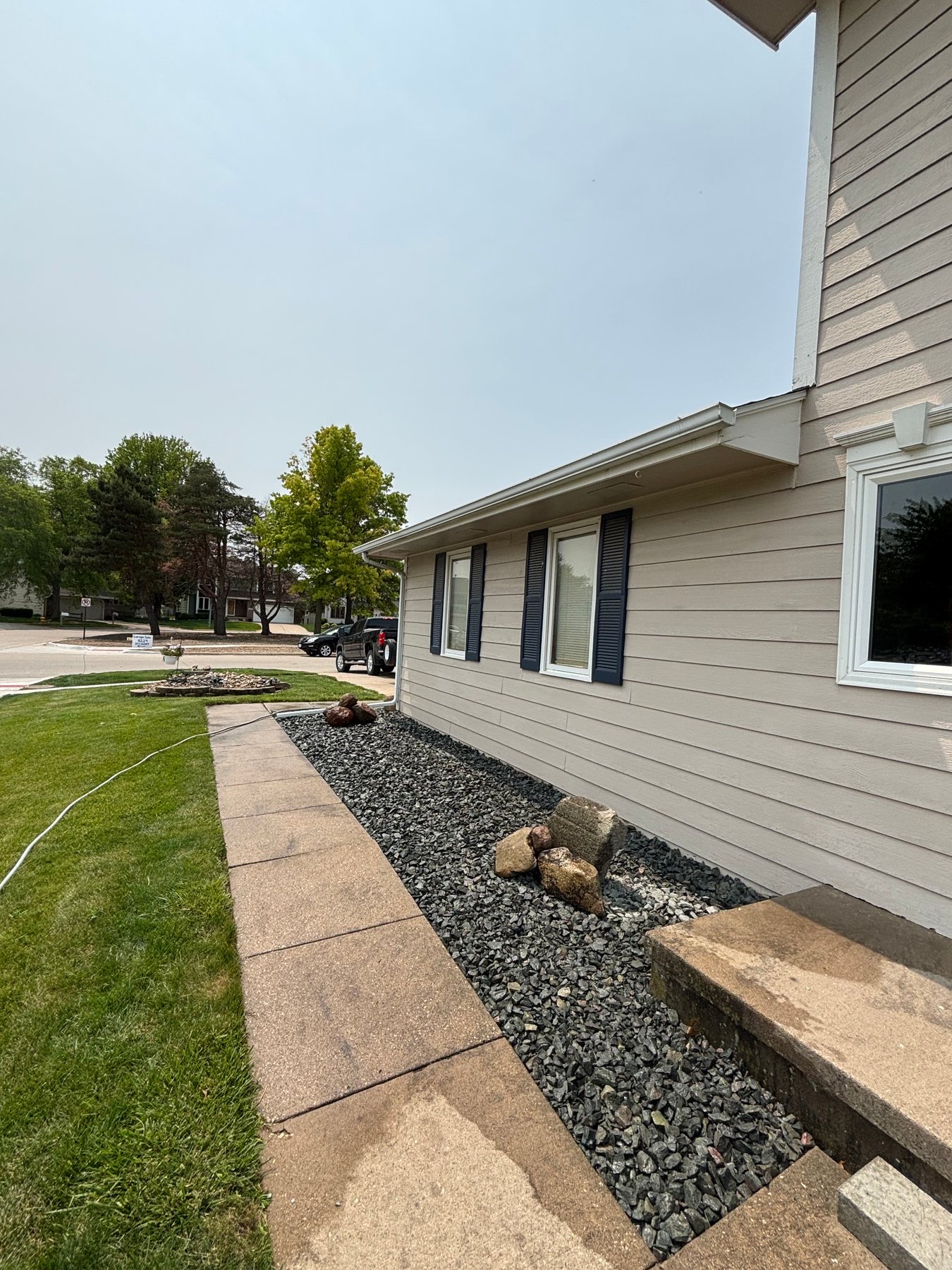 A landscaped bed with dark grey gravel and decorative boulders alongside a house with light tan siding and a concrete walkway.