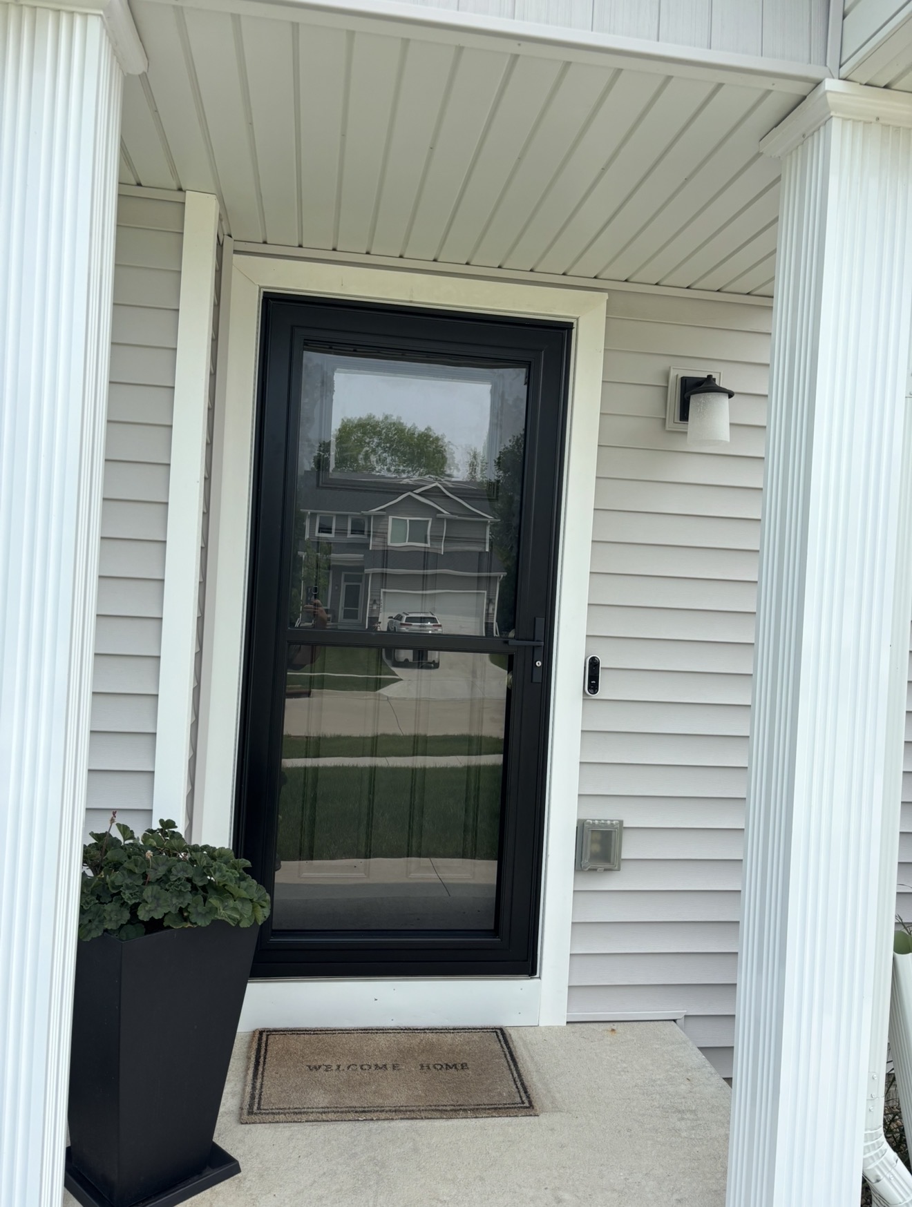 A black-framed storm door installed on a residential entryway with white trim, light grey siding, and fluted columns.