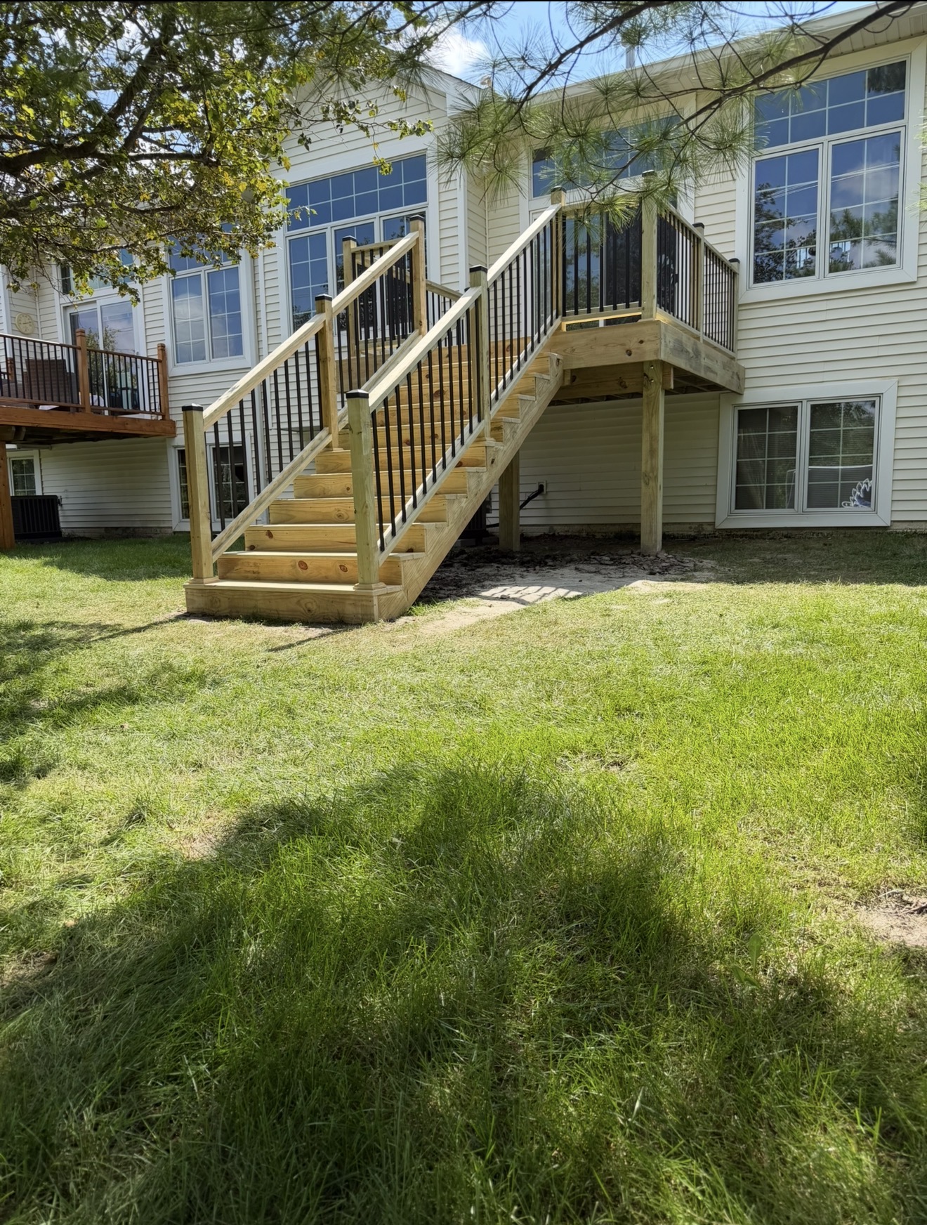 Newly constructed pressure-treated wood deck stairs with black metal spindles leading down to a grassy backyard from a beige house.