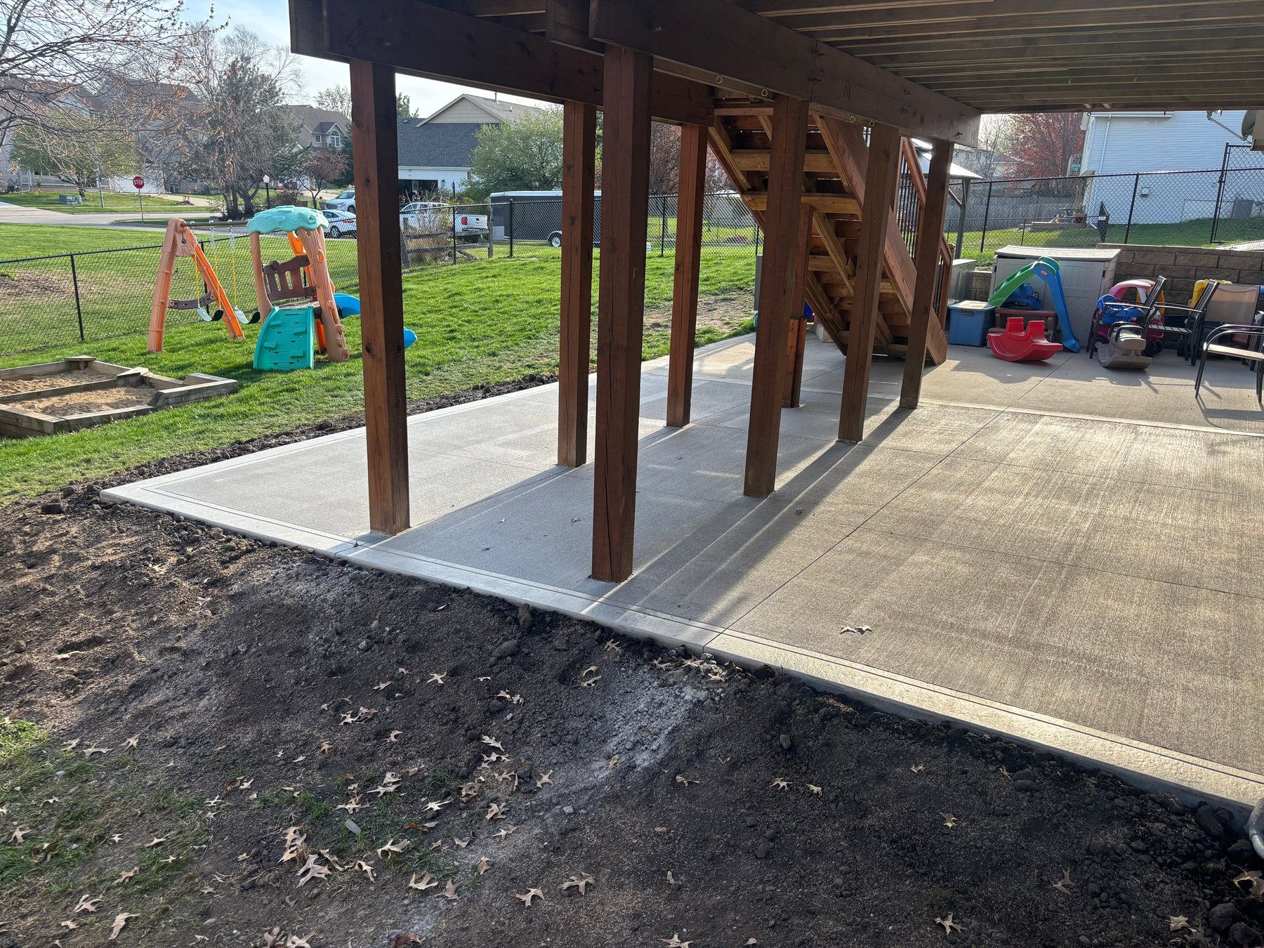 A newly poured concrete patio with a brushed finish sits beneath the dark stained wooden support posts of an elevated deck.