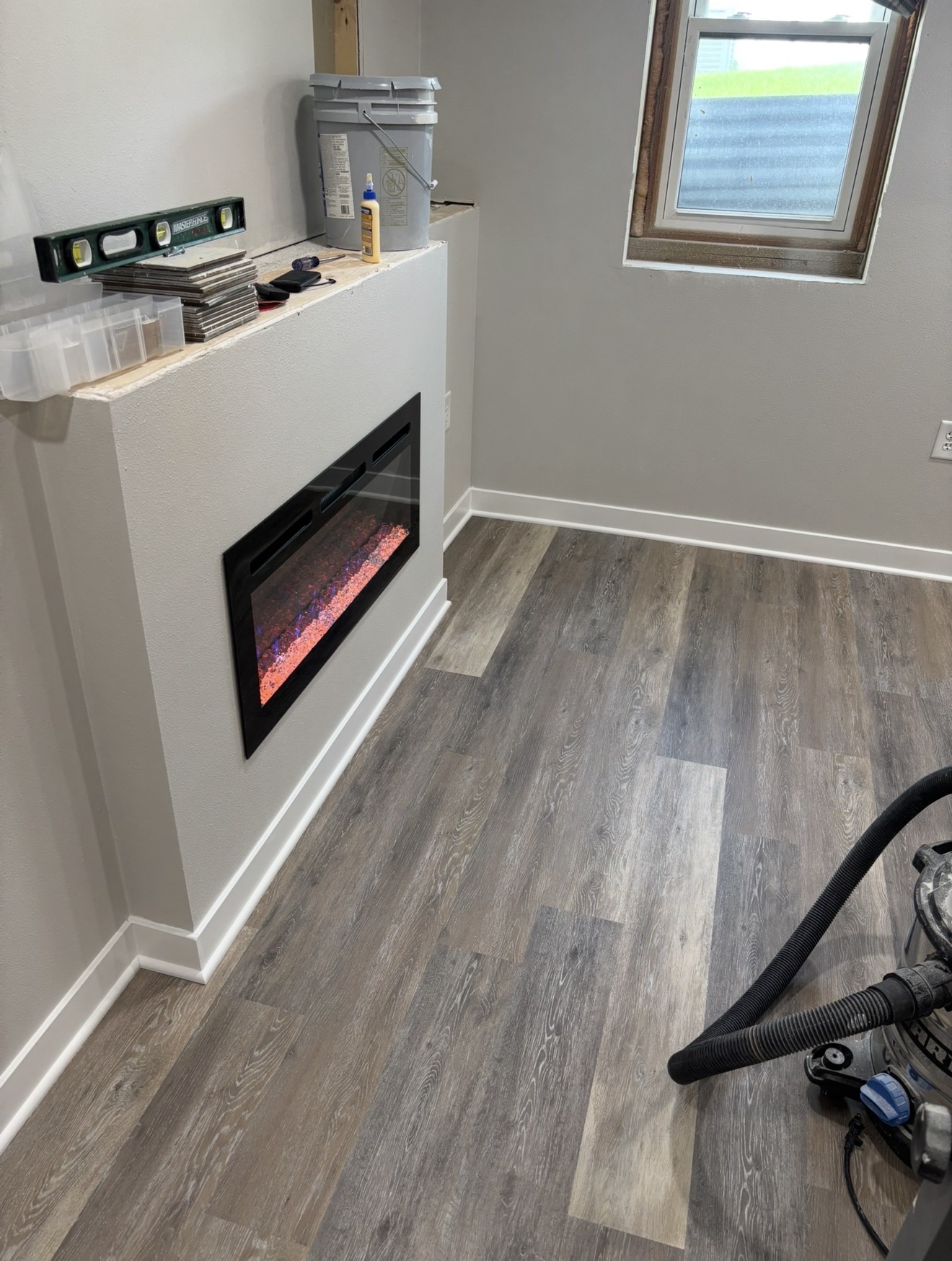 A basement room under construction featuring gray wood-look plank flooring, light gray walls with white baseboards, and an inset electric fireplace.