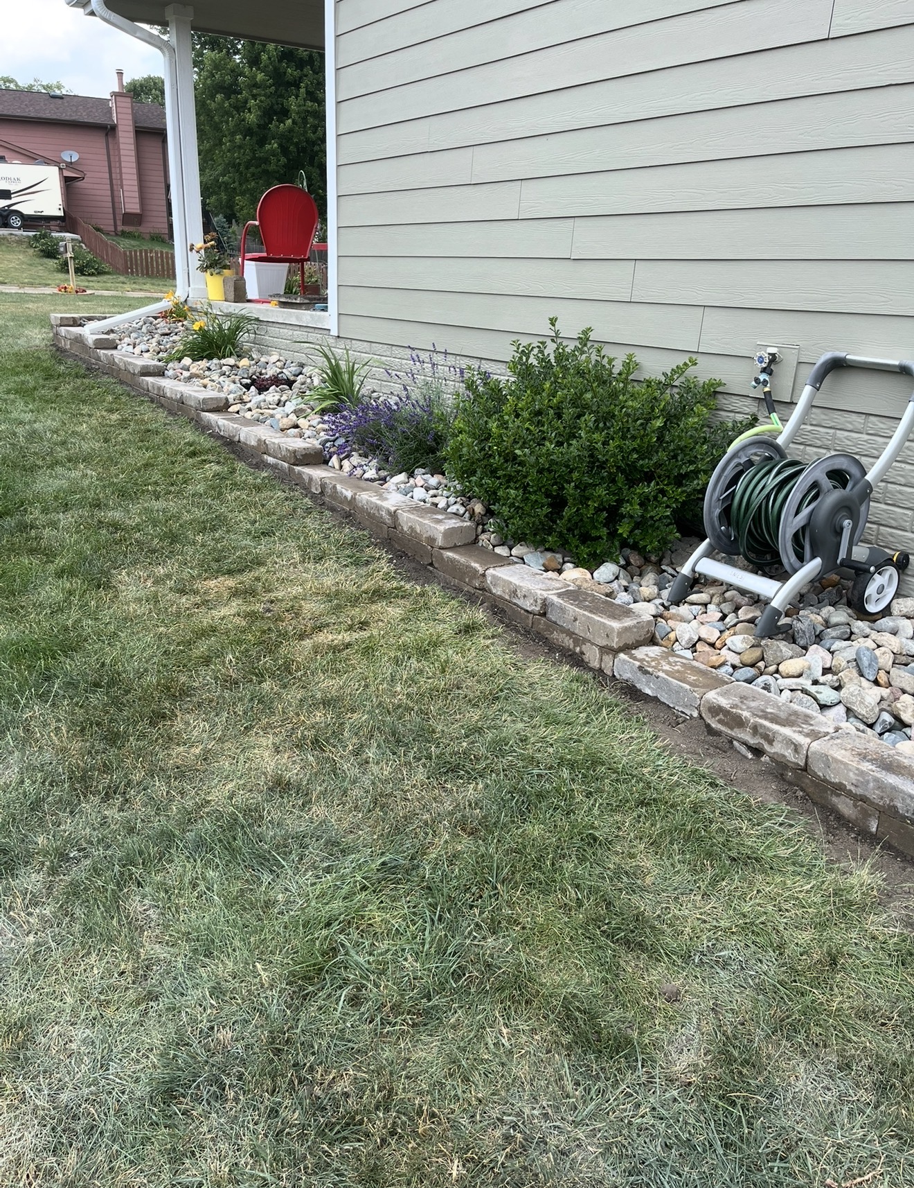 A newly installed landscape garden bed alongside a house with a stone block border, river rocks, plants, and shrubs next to green siding.