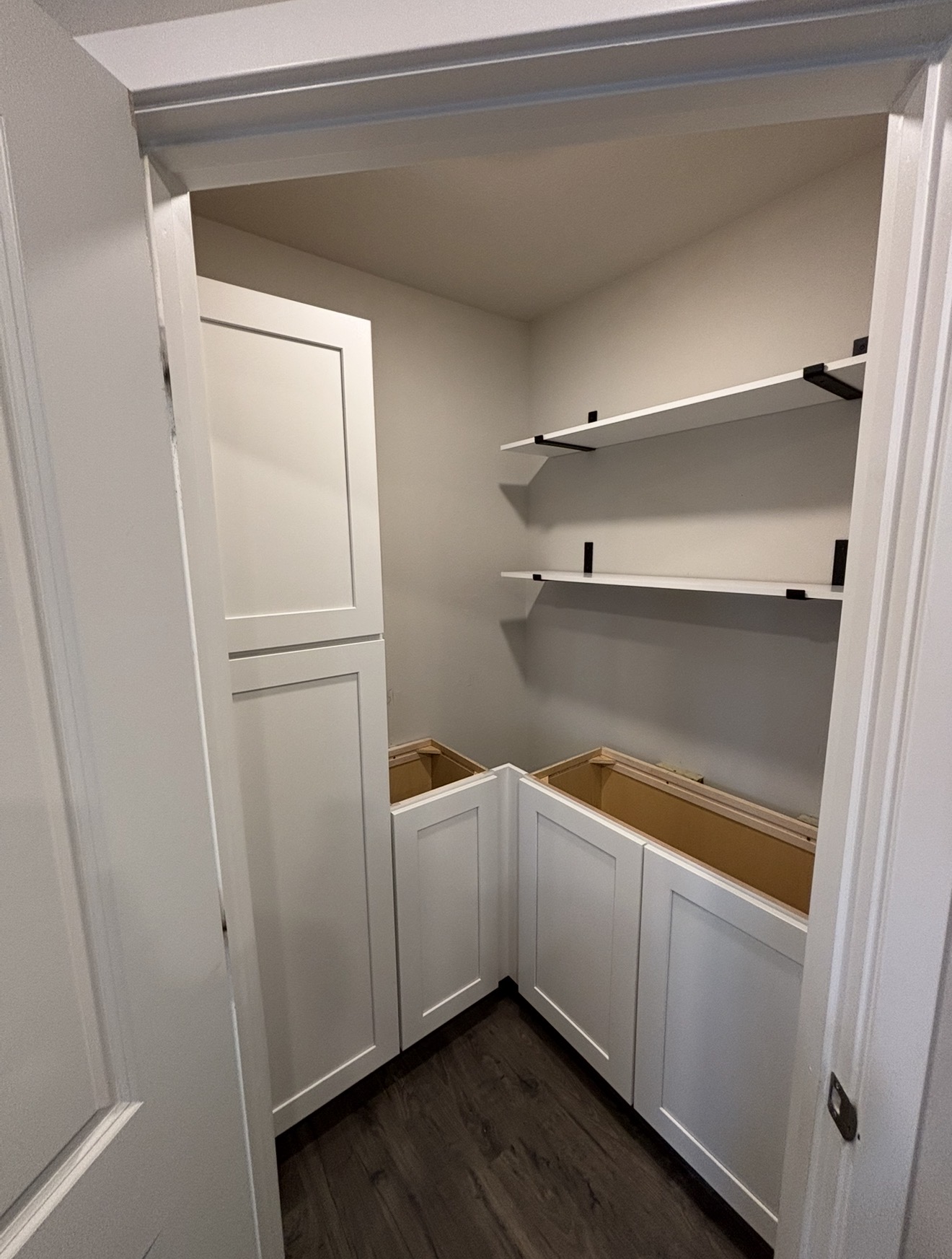 A walk-in pantry featuring white shaker cabinets, floating white shelves with black brackets, and dark wood flooring.