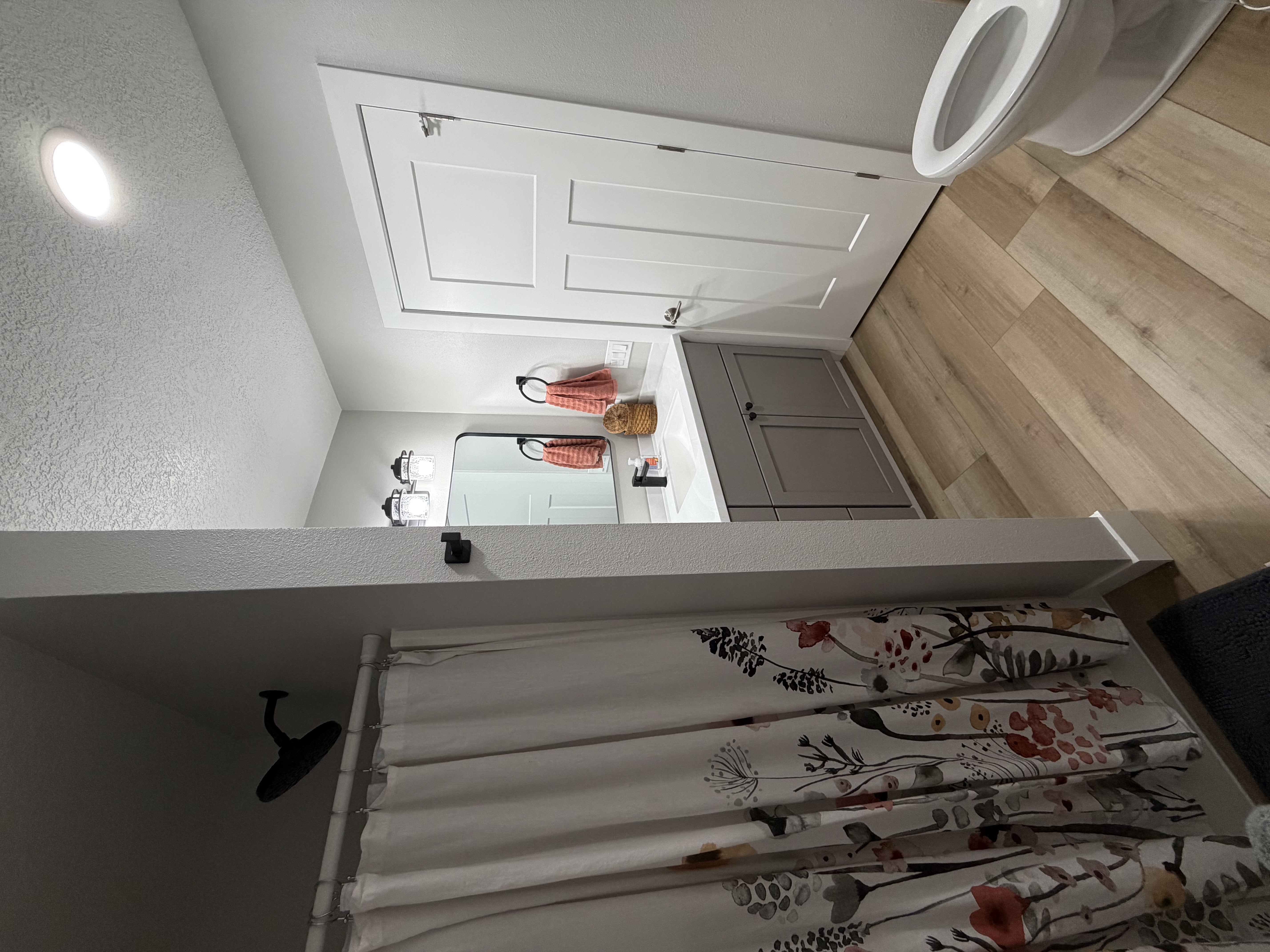 A modern bathroom featuring light wood flooring, a grey vanity with a white countertop, a white panel door, and a floral-patterned shower curtain.