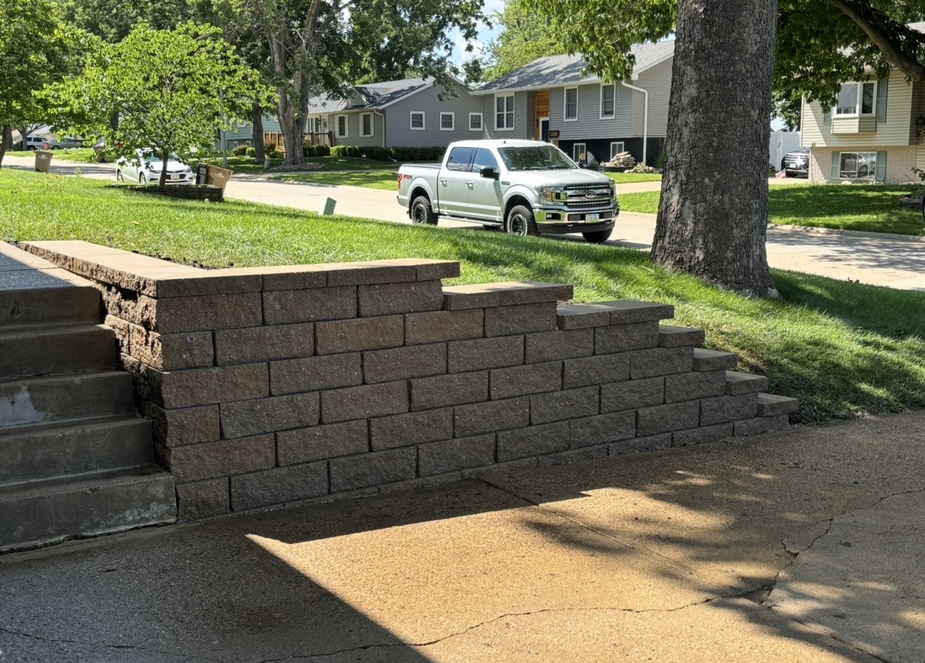 A tiered retaining wall constructed from brown textured concrete blocks next to a grassy slope and driveway.