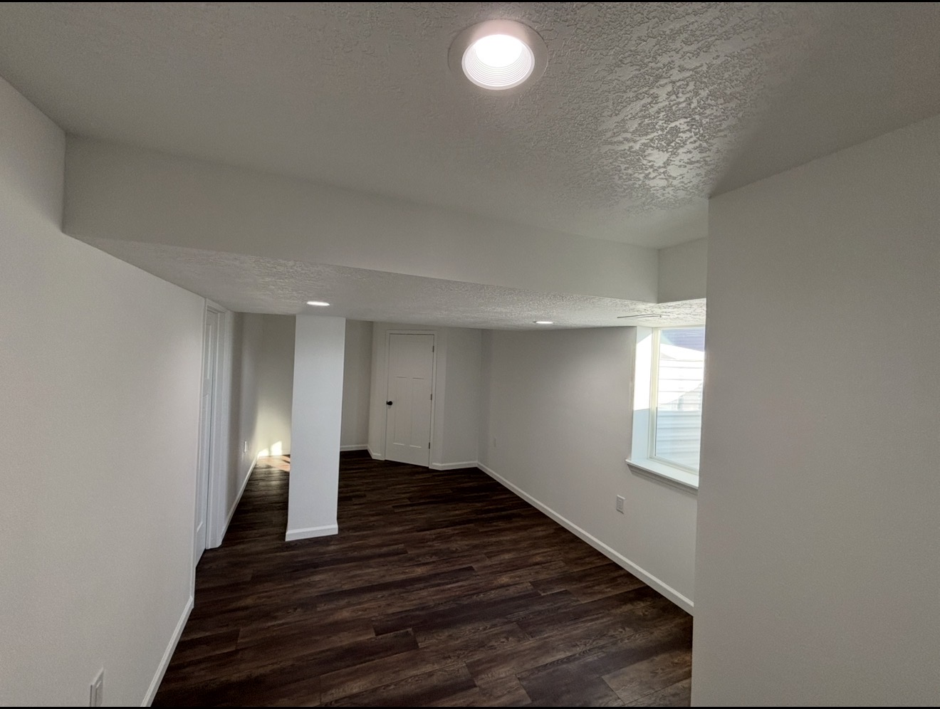 A finished basement featuring dark wood flooring, light grey walls, a white support column, and recessed ceiling lighting.