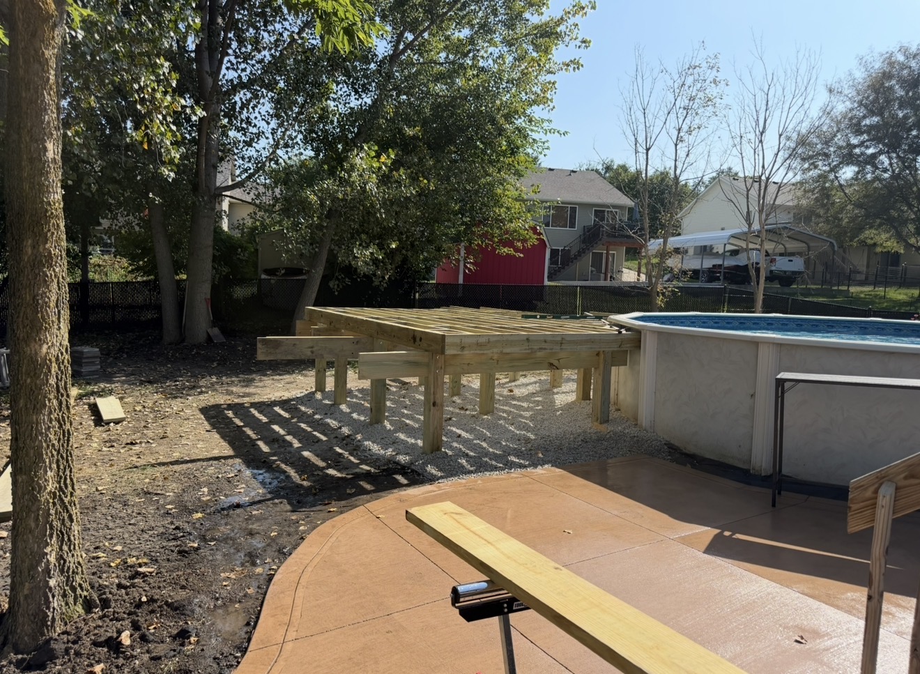 Unfinished wood framing for a backyard pool deck next to an above-ground swimming pool and a concrete patio.