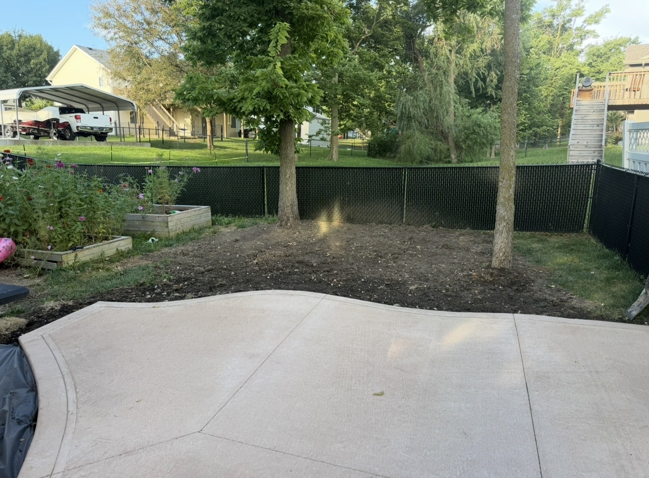 A curved concrete patio with a decorative border next to a freshly graded dirt yard area and a black privacy fence.