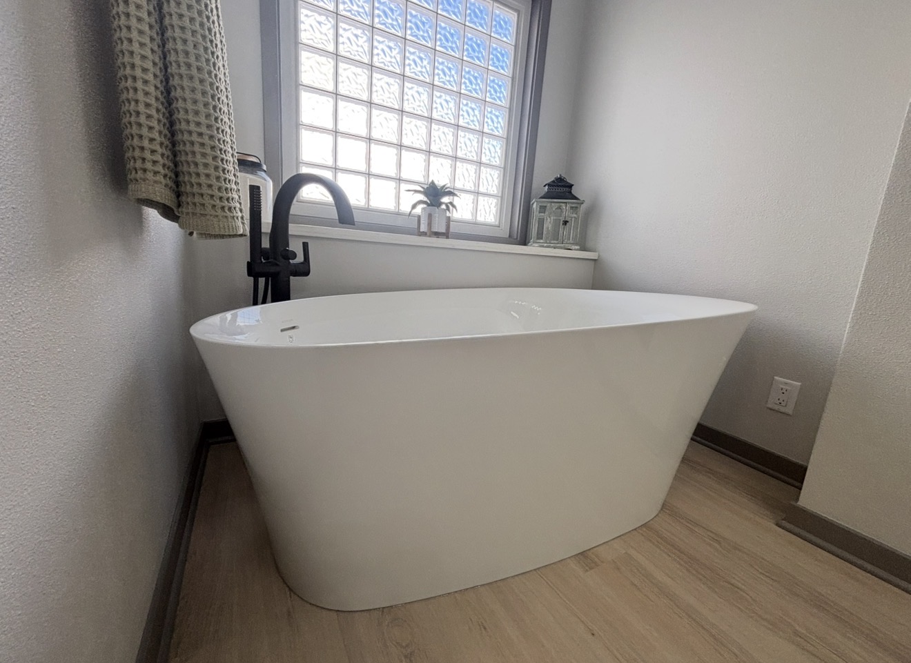 A modern white freestanding bathtub on light wood flooring, featuring a matte black floor-mounted faucet in front of a glass block window.