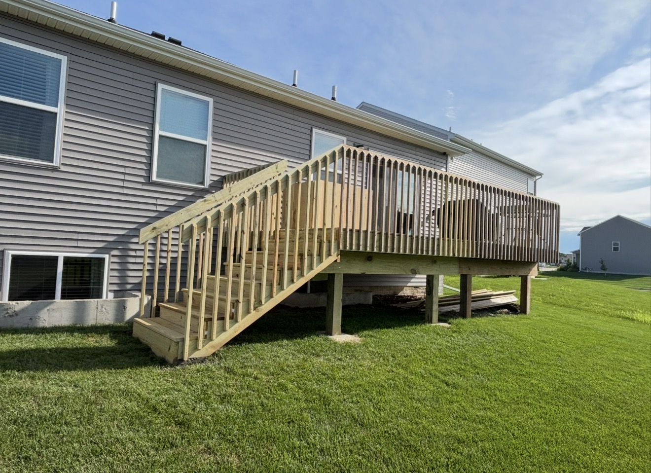 A newly constructed pressure-treated wood deck with stairs and vertical balusters attached to a grey vinyl-sided house.