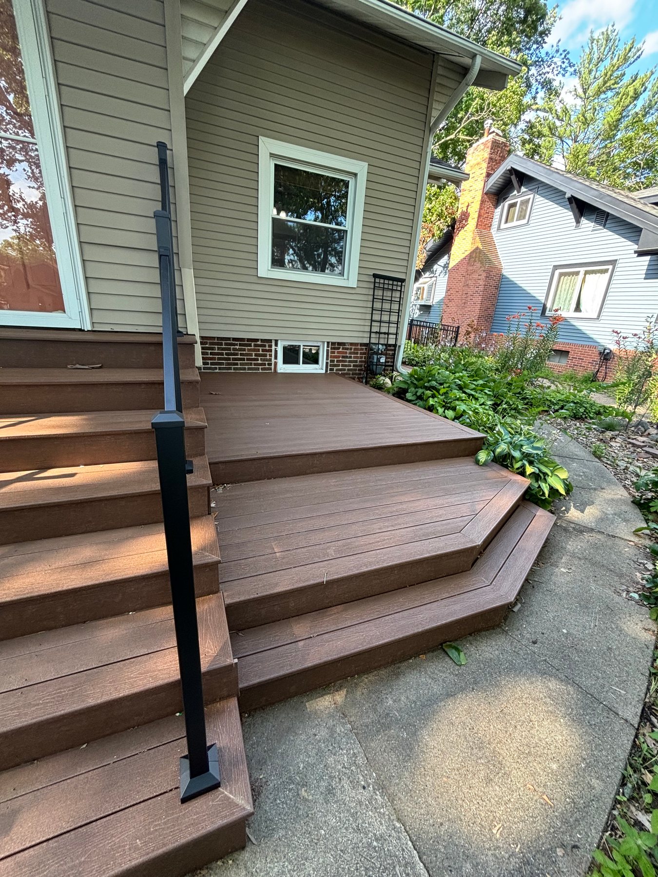 Multi-level brown composite deck stairs with a black metal handrail leading to a concrete walkway.