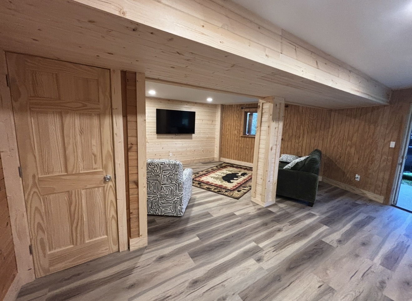 Finished basement living area with light pine wood paneling, a matching solid wood door, grey wood-look vinyl flooring, and a wood-wrapped support beam and column.