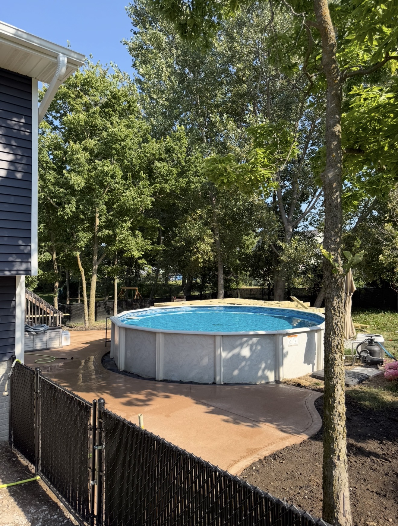 An above-ground swimming pool surrounded by a custom curved tan concrete patio and a black chain-link fence.