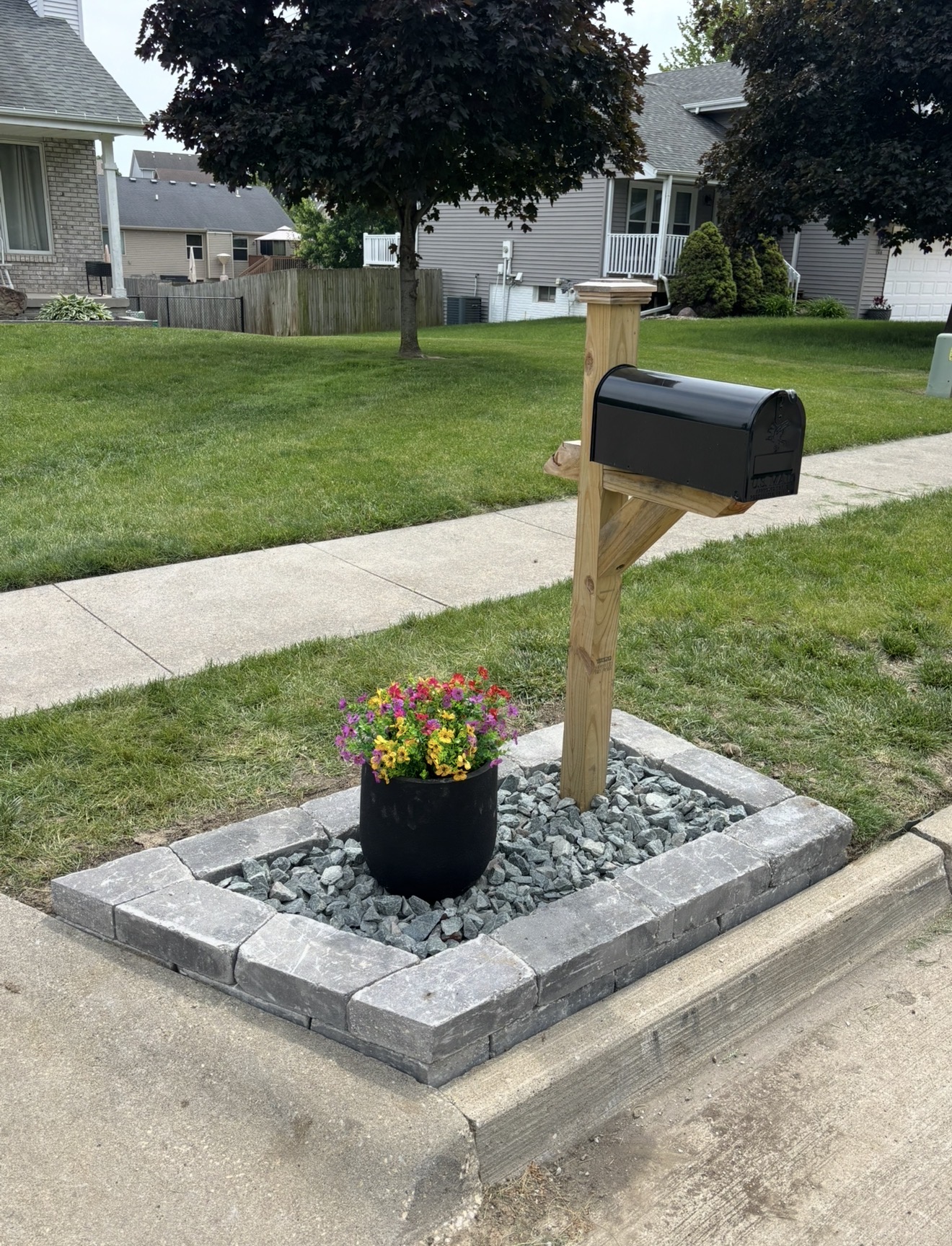 A new wooden mailbox post with a black mailbox, surrounded by a grey stone block border filled with decorative gravel and a colorful potted flower.
