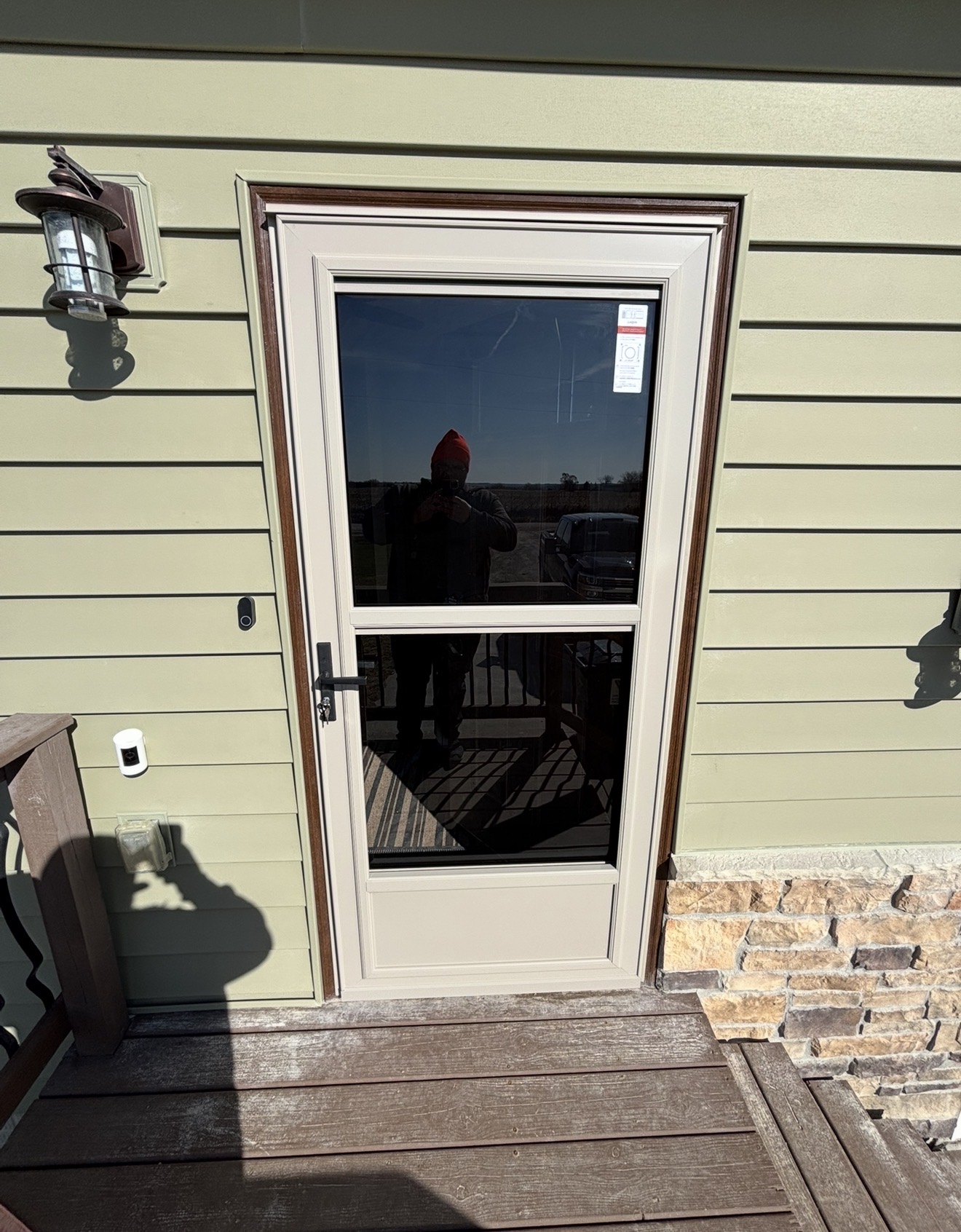 A beige storm door with dark brown trim installed on a house with light green siding and stone veneer.