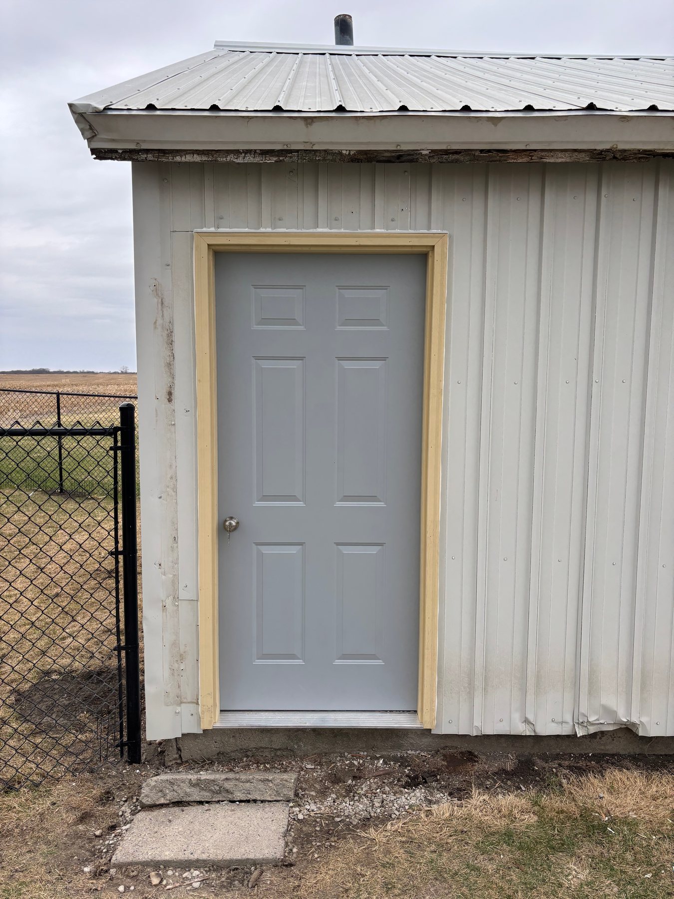 A grey six-panel exterior door with a silver handle and light wood trim installed on a white metal-sided shed.