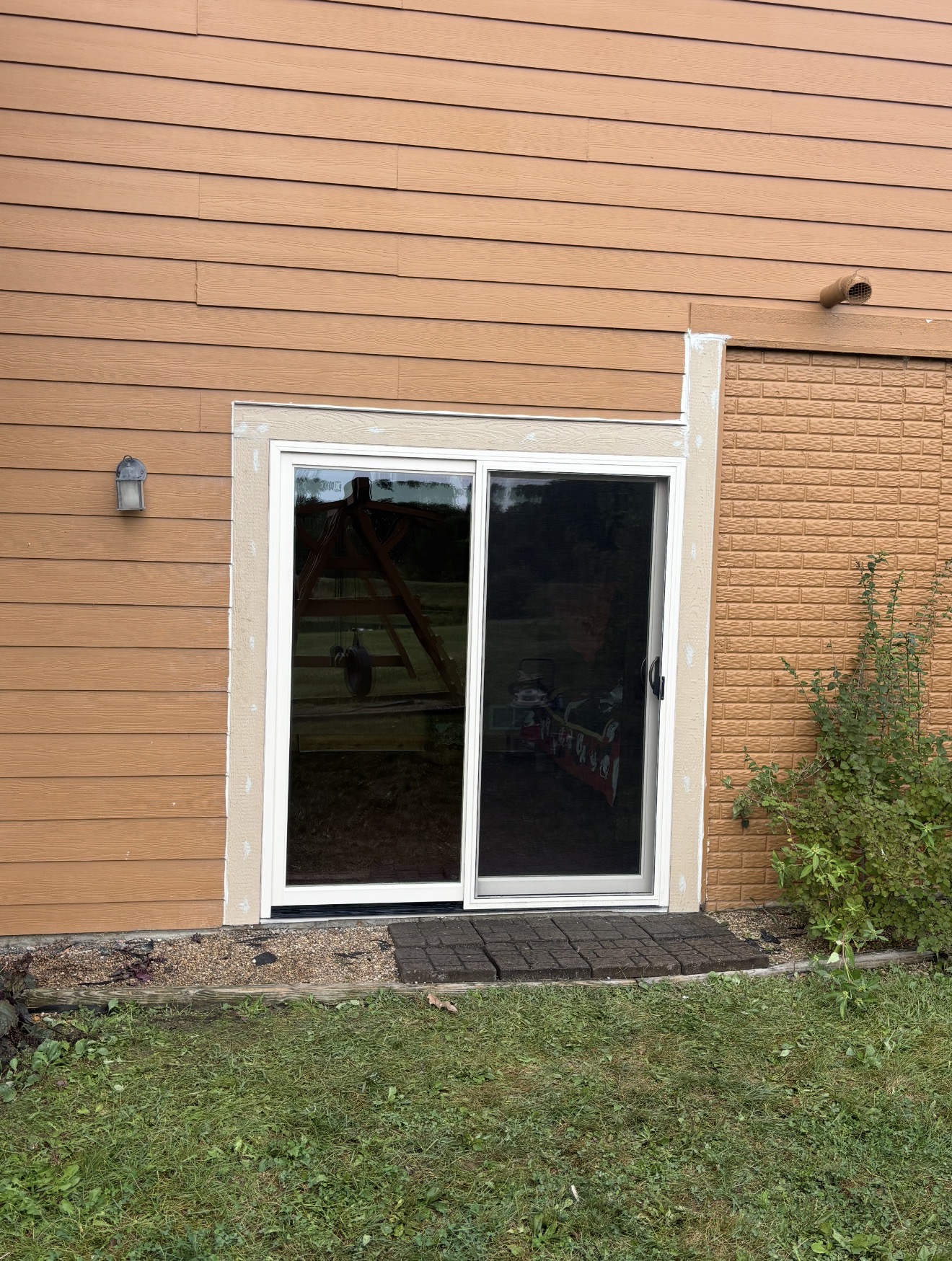 A newly installed white sliding glass patio door set into brown horizontal siding with unfinished light trim.