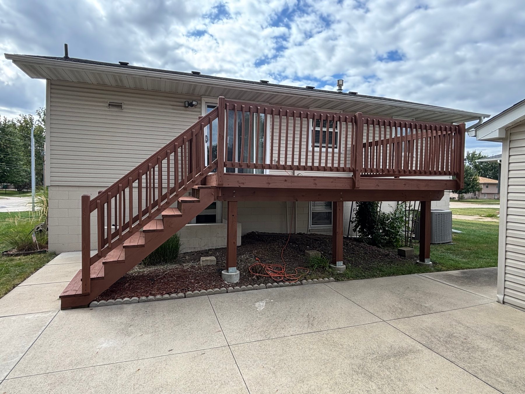 An elevated wooden deck and stairs stained in a solid reddish-brown color, attached to a house with beige siding.
