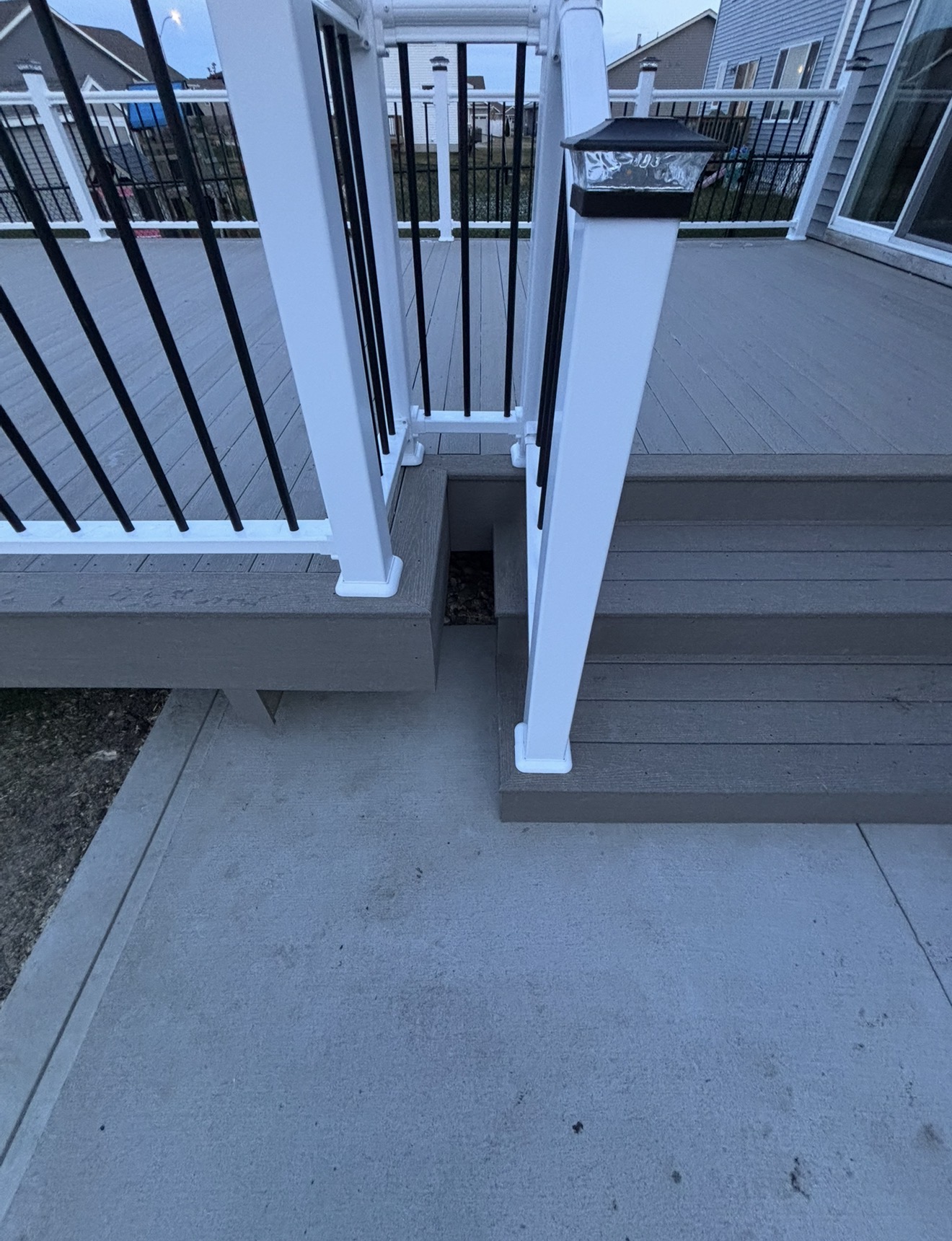 Grey composite deck stairs with white railing posts, black balusters, and a post cap light leading down to a concrete patio.