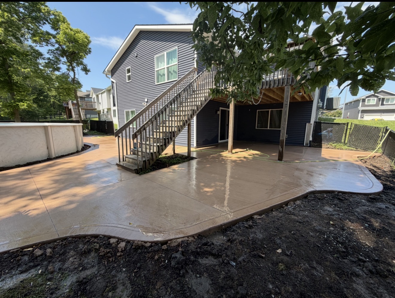 A freshly poured, tan-stained concrete patio with curved borders sits beneath a wooden deck and staircase behind a house with dark siding.