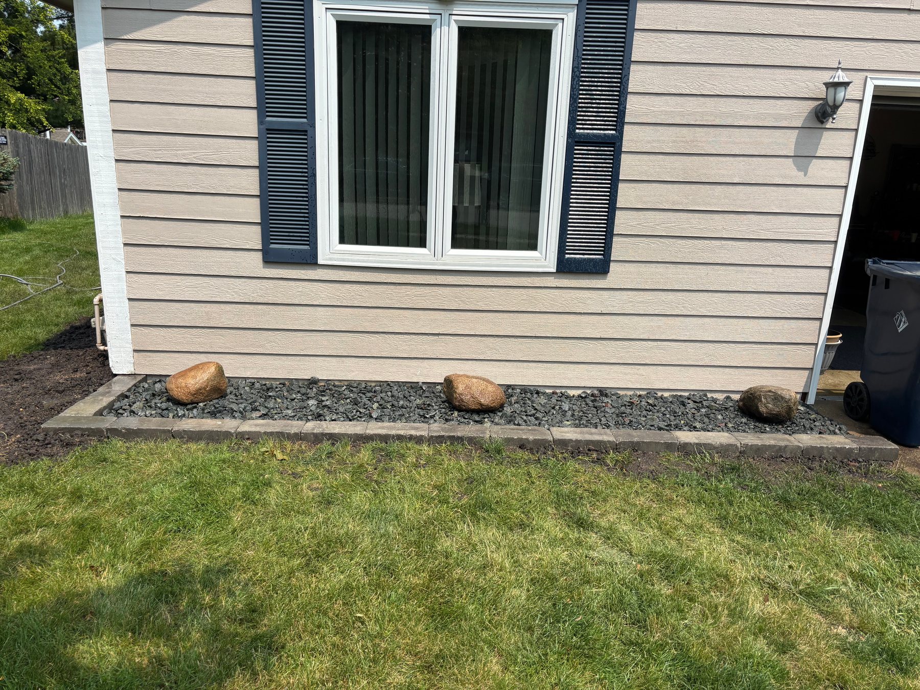 Landscape garden bed along tan house siding featuring dark gray stone borders, black gravel mulch, and three decorative brown boulders.