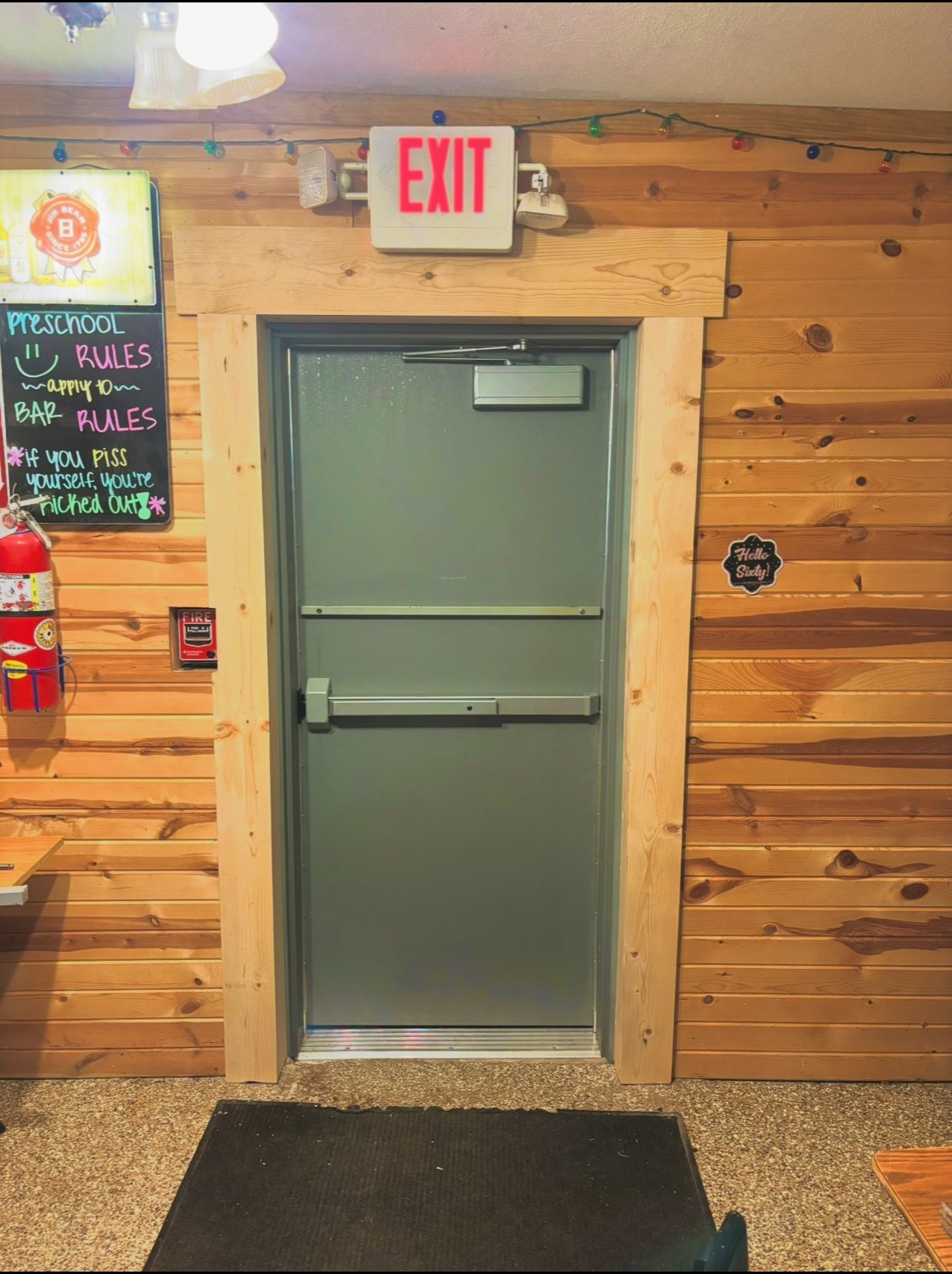 A grey metal exit door with a push-bar handle, framed by light-colored natural wood trim against a horizontal wood-paneled wall.