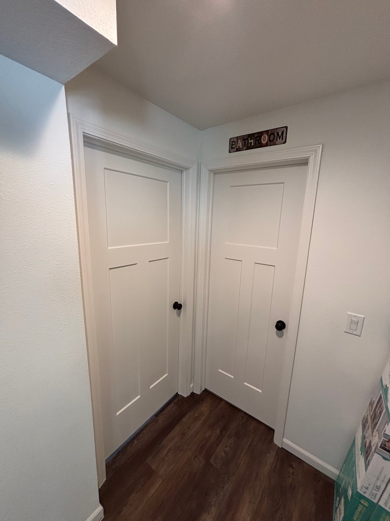 Two white three-panel craftsman-style interior doors with black knobs and white trim set against dark wood flooring.
