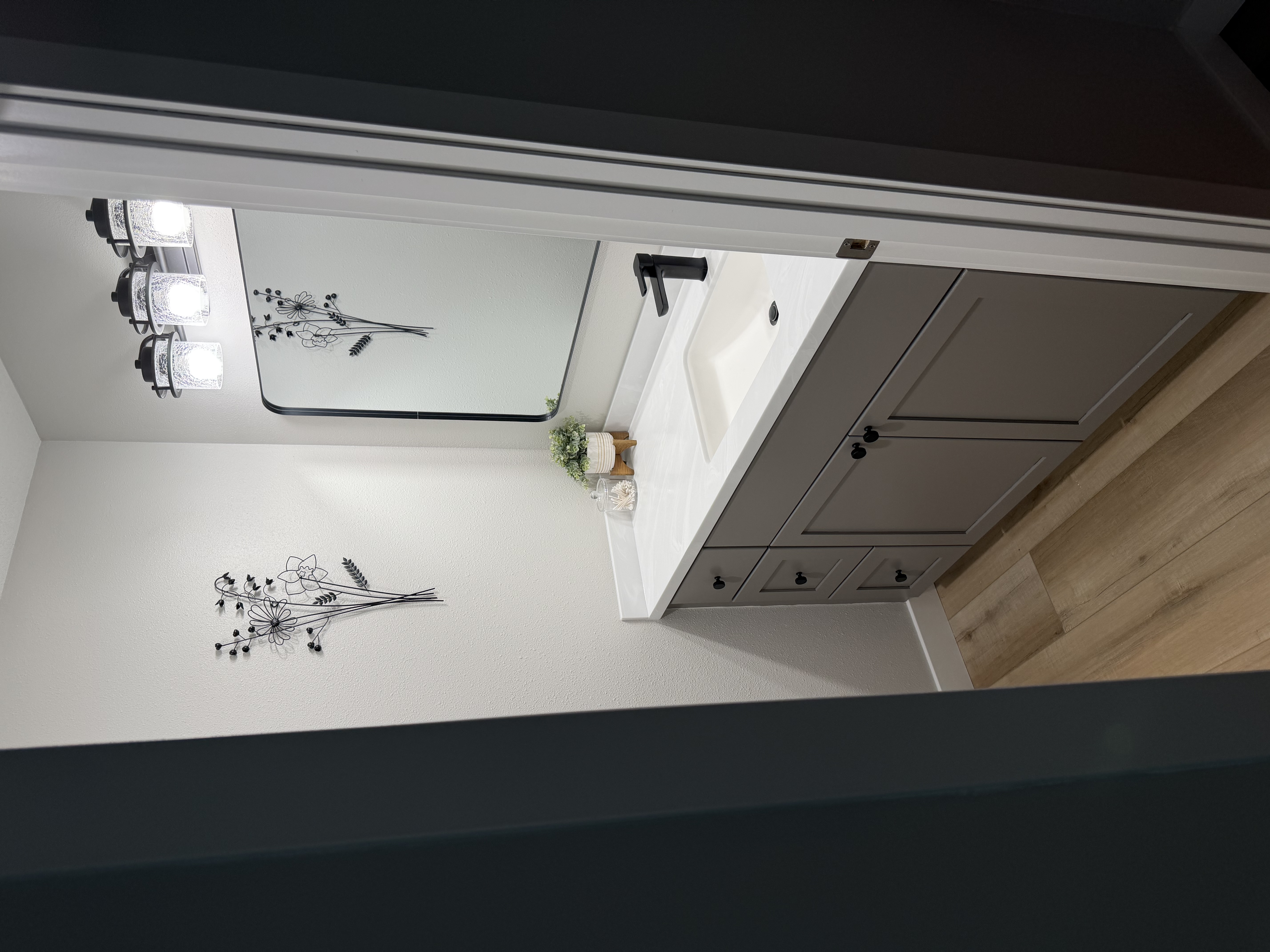 A modern bathroom vanity featuring gray cabinets, a white countertop, black hardware, a matching black-framed mirror, and light wood-look flooring.