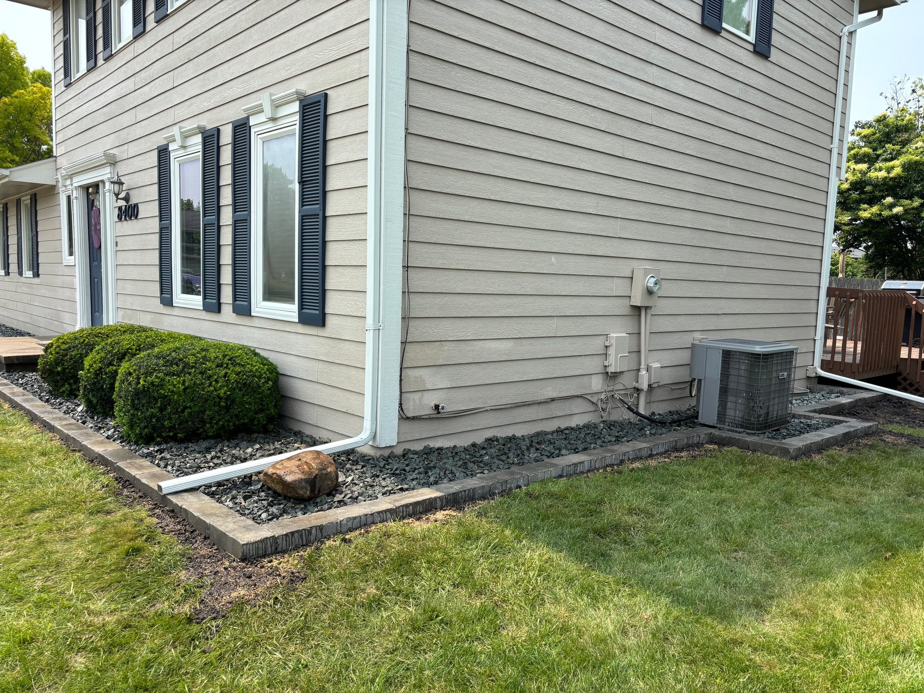 A stone-bordered landscaping bed filled with dark gray gravel and green shrubs runs along the foundation of a beige two-story home.