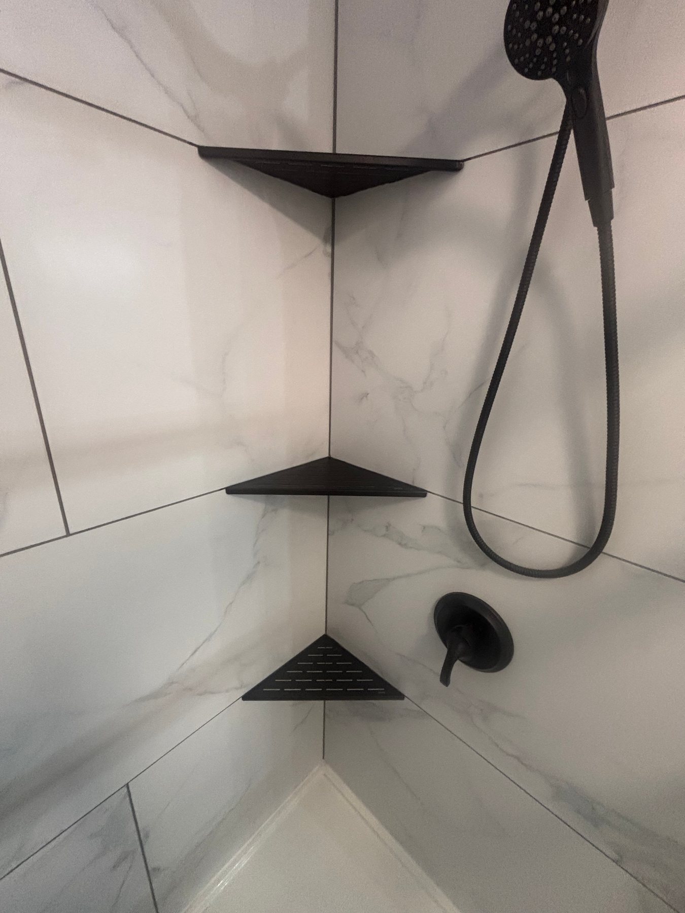 White marble-look tiled shower corner with three matte black triangular shelves and matte black shower fixtures.