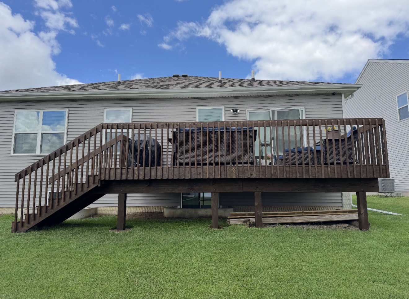 An elevated dark brown stained wooden deck with stairs attached to the back of a light gray vinyl-sided house.