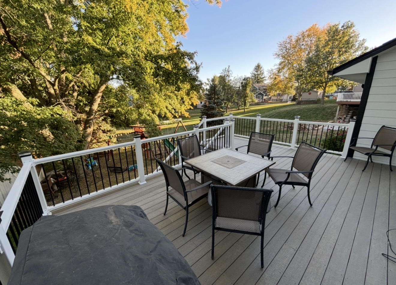 An elevated composite deck with light gray planks, white railings with black spindles, and a patio dining set with five chairs around a square table.