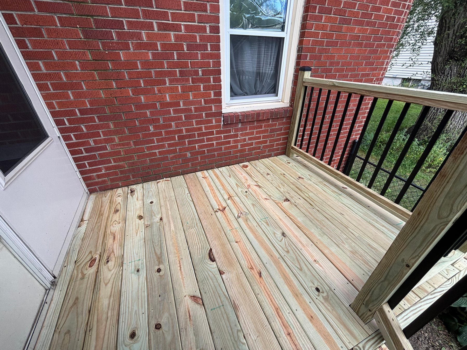A newly built wooden deck with pressure-treated pine boards and a railing featuring black metal balusters, set against a red brick house wall.