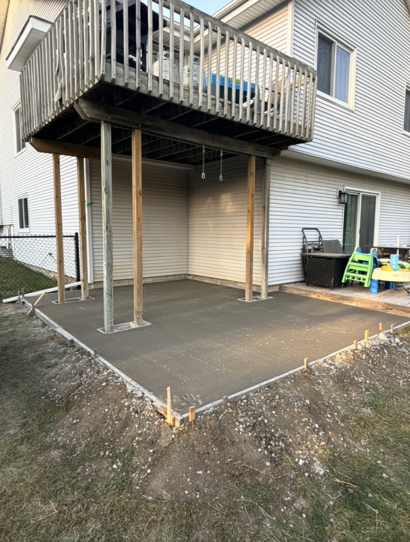 A freshly poured concrete patio slab under construction beneath an elevated wooden deck with support posts.