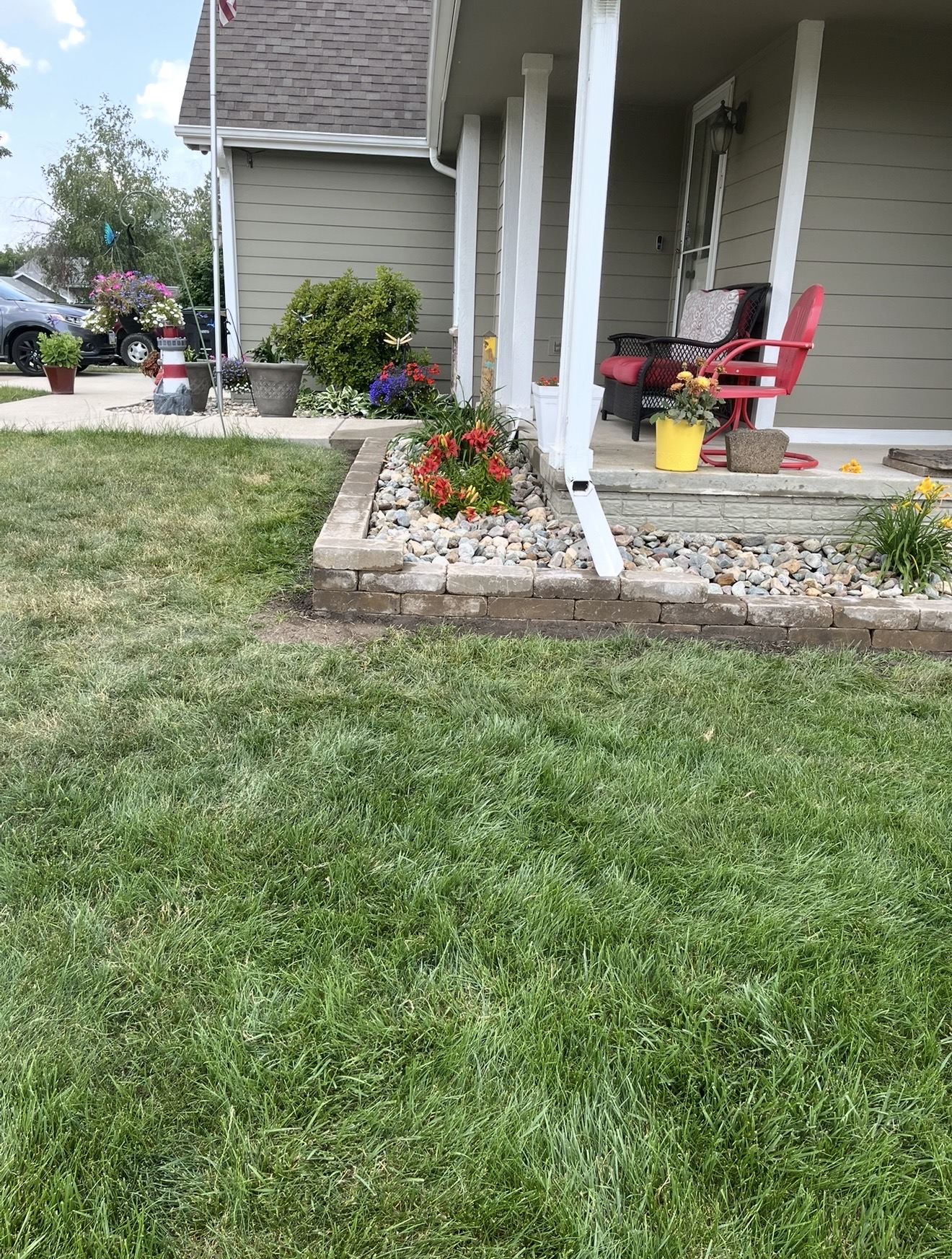 A stone retaining wall border filled with river rocks and flowers sits next to a green grass lawn and front porch.