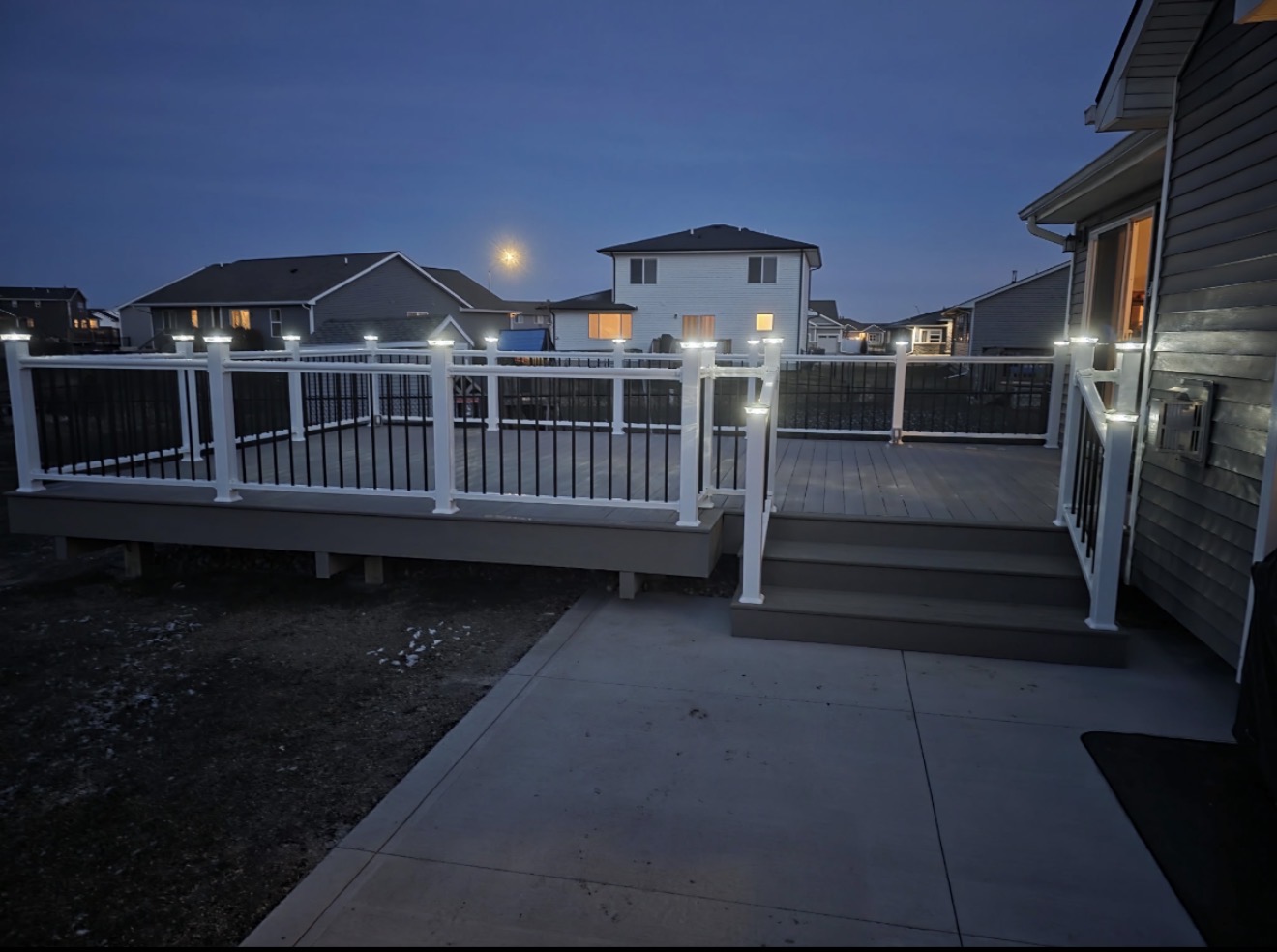 A gray composite deck at dusk featuring white railings with black balusters and illuminated post caps, with steps leading down to a concrete patio.
